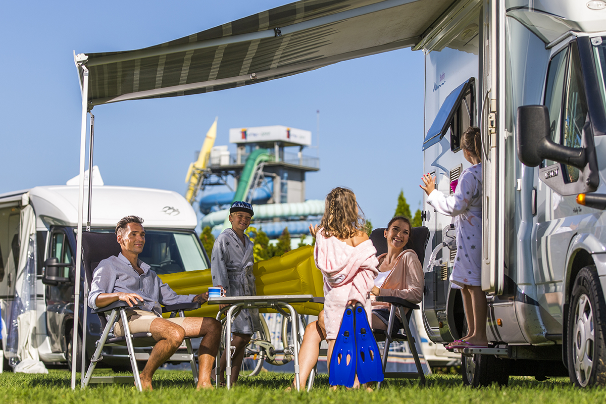 Pet ljudi stoji in sedi za mizo pod baldehinom avtodoma in se pogovarja