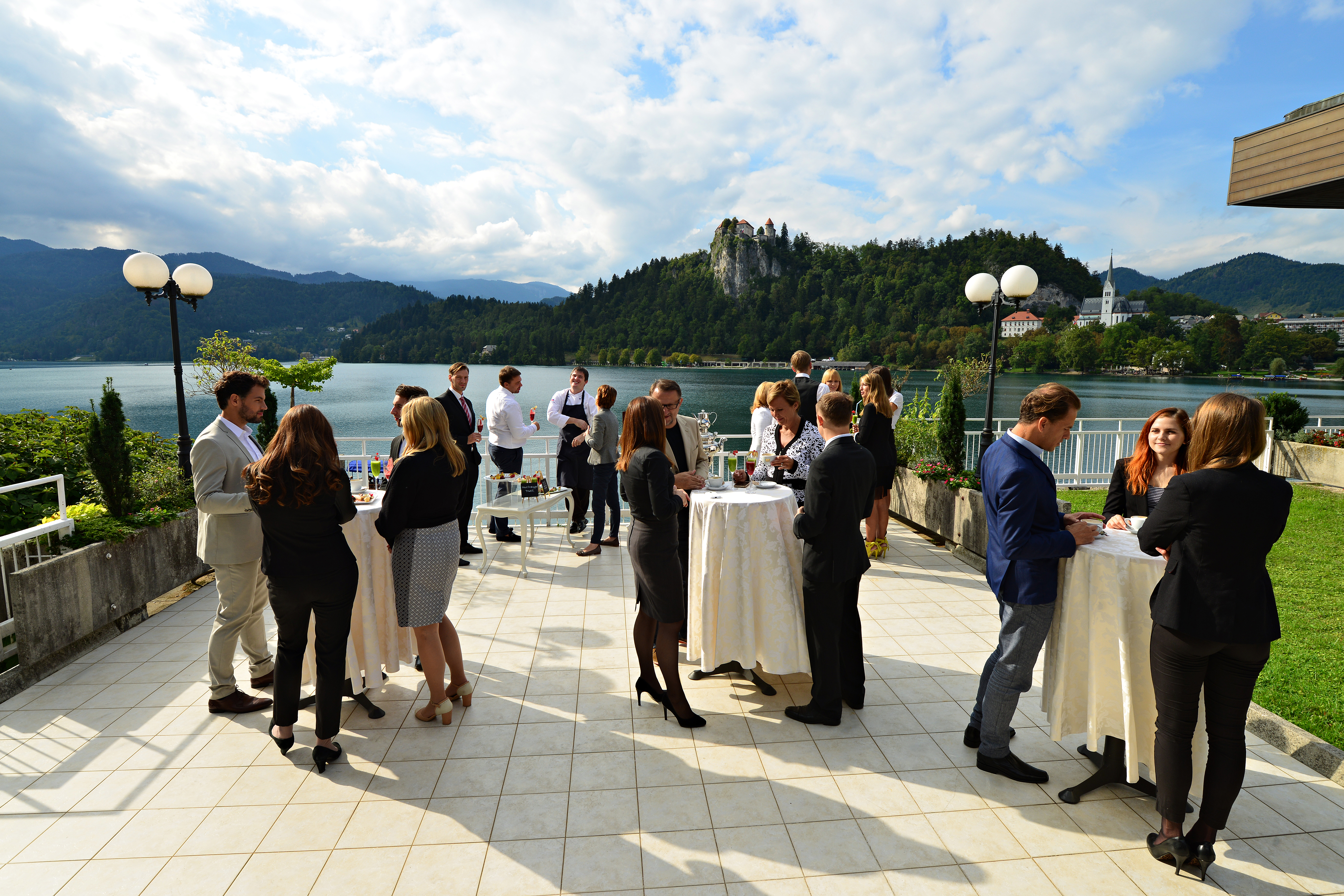 Skupina poslovnežev se na ploščadi pri blejskem jezeru druži ob visokih mizah in buffetu