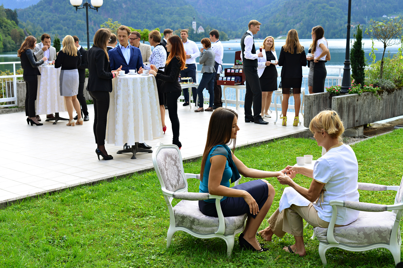 Skupina ljudi v poslovnih oblekah se druži ob visokih mizah na terasi pri jezeru