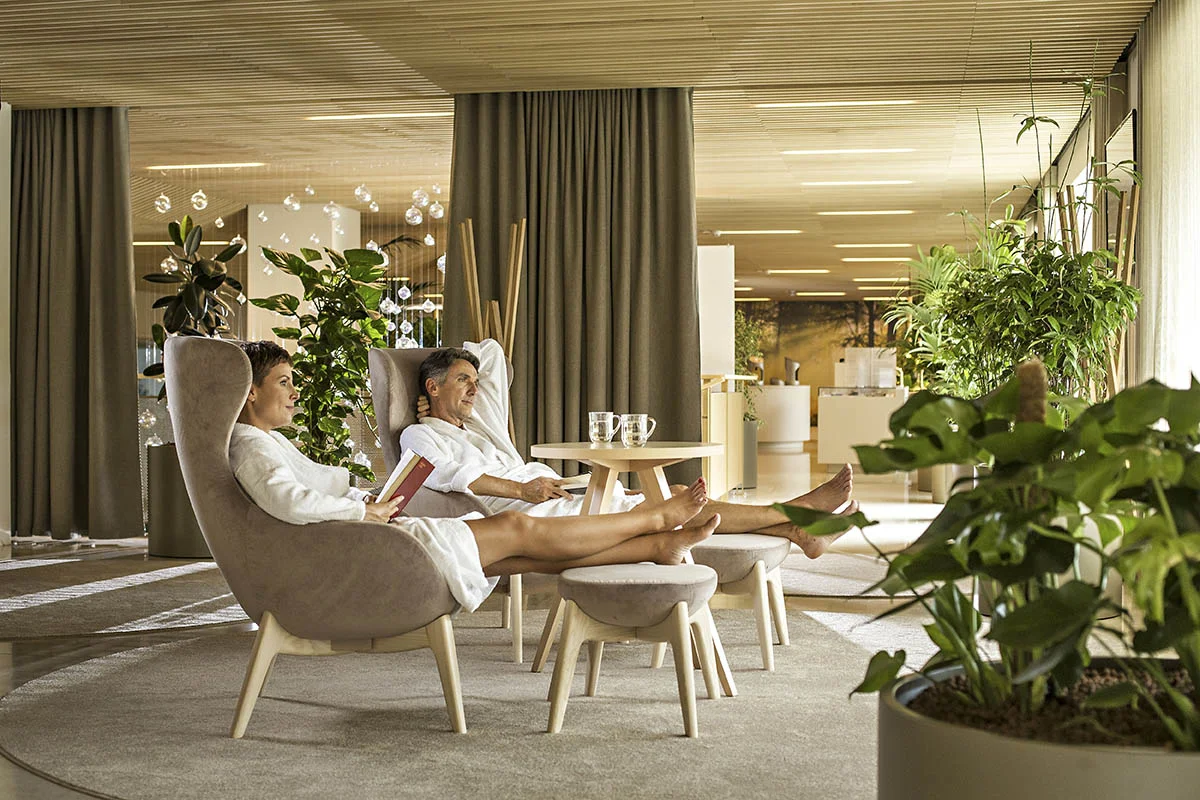 Moški in ženska v belih haljah počivata na ležalniku v zelenem prostoru termalnega parka s knjigo v roki