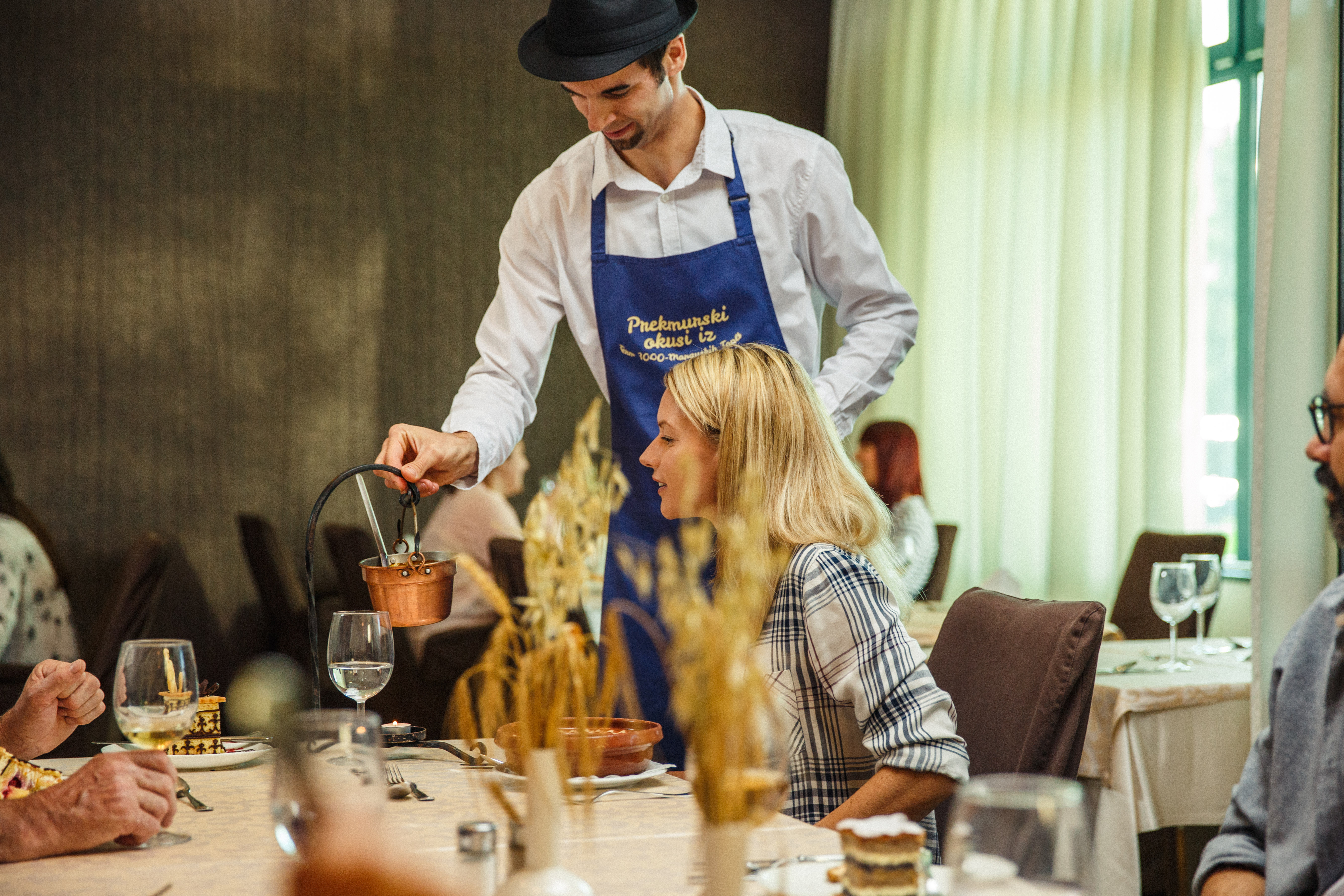 Ženska sedi za hotelsko mizo, natakar z modrim predpasnikom pa ji razlaga prekmursko jed