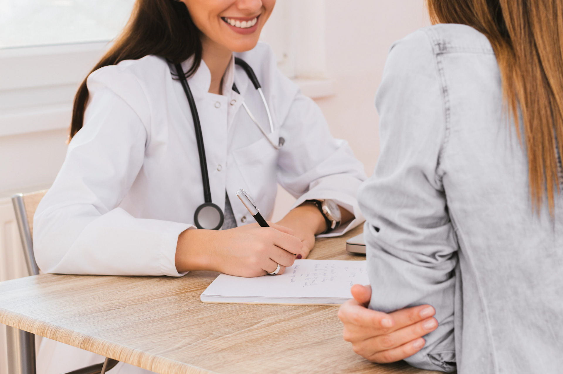 Nasmejana zdravnica v beli halji sprašuje pacientko in si zapisuje ugotovitve